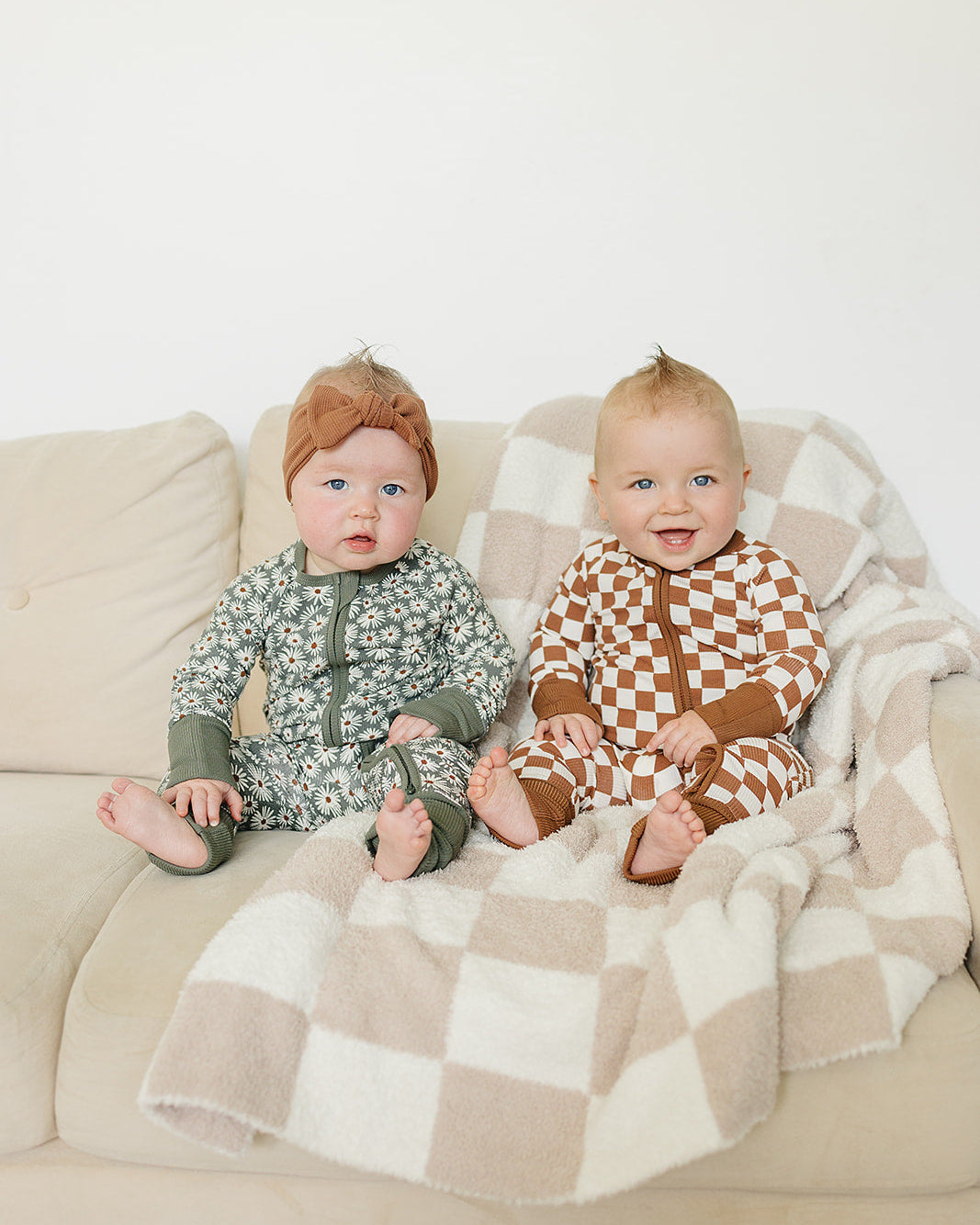 Two babies sitting on a couch with a checkered blanket