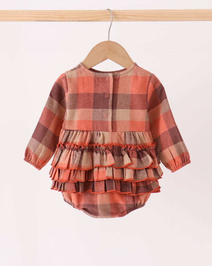 A baby long-sleeve romper with a warm-toned plaid pattern in orange, rust, and beige, featuring a tiered ruffle hem and button front, hanging on a wooden hanger.