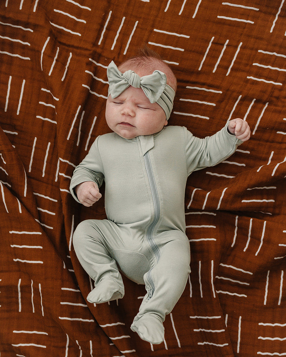 Newborn baby in a light gray outfit with a bow on a brown patterned blanket