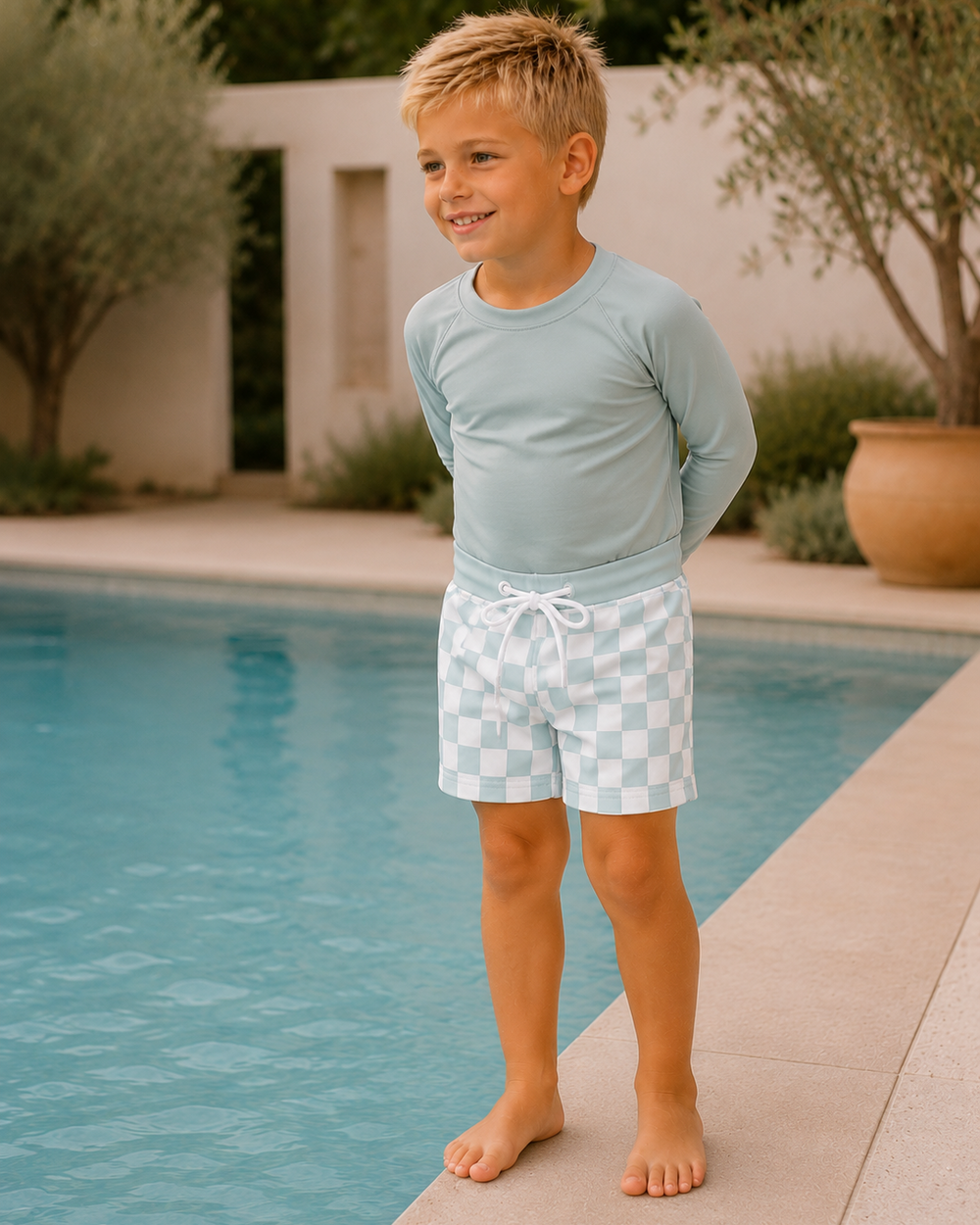 Young boy in light blue shirt and checkered shorts standing by a pool.