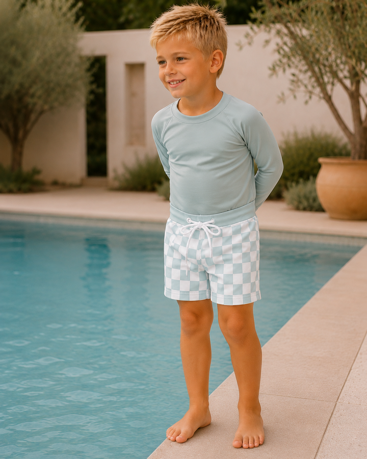 Young boy in light blue shirt and checkered shorts standing by a pool.