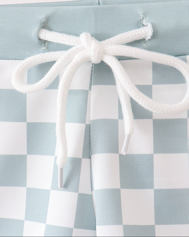 Checkered swim trunks with white tieback on a light blue and white checkered fabric background.