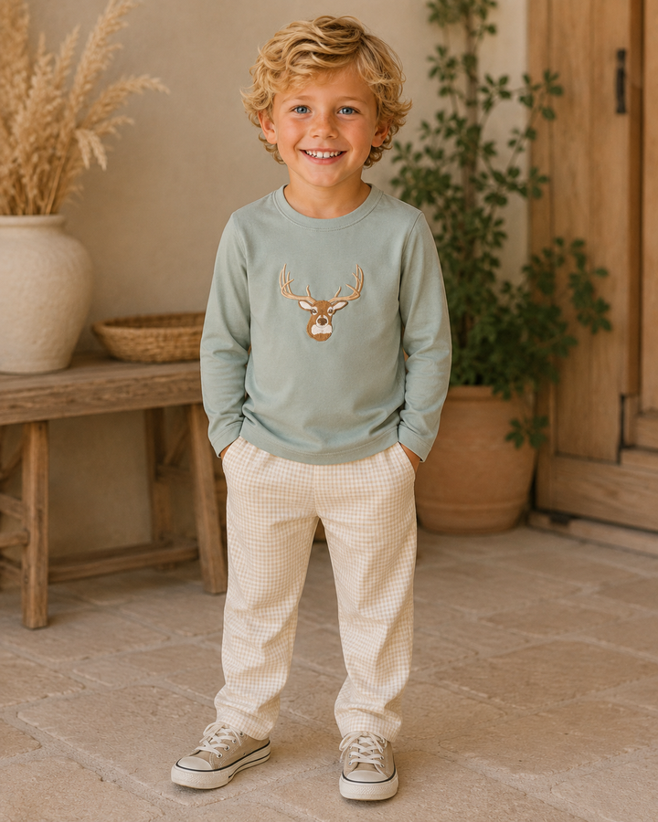 A young boy with curly blonde hair stands smiling in a rustic indoor setting, wearing a sage green long-sleeve shirt with a deer graphic on the chest, cream gingham pants, and canvas sneakers.