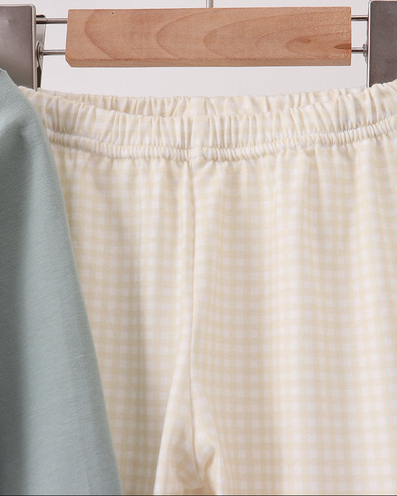 Close-up of a pair of cream and white gingham check lounge pants hanging on a wooden hanger, with a soft sage green fabric partially visible on the left.