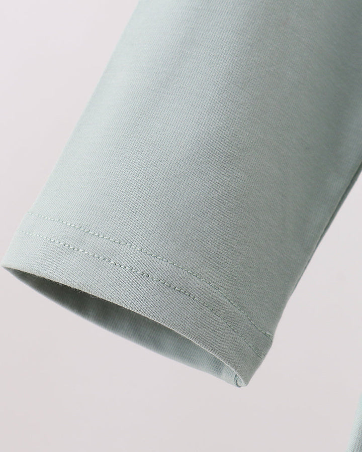 Close-up detail of a light sage green sleeve showing the hem and fine stitching against a pale pink background.