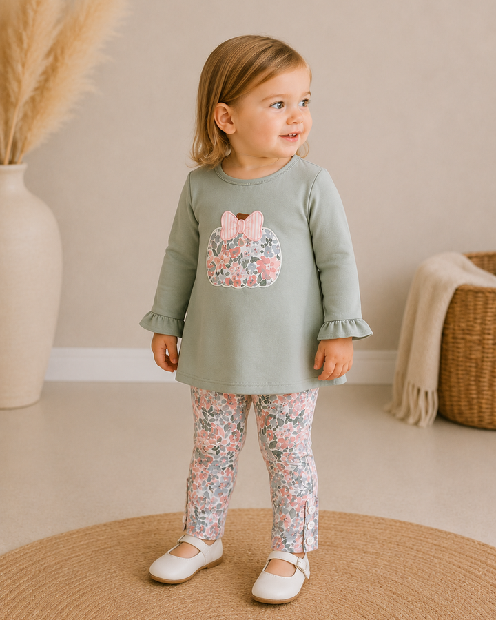 A toddler girl wearing a sage green long-sleeve tunic top with a pink floral bunny applique on the chest, paired with matching floral leggings and white shoes, standing on a round woven rug in a softly decorated room.