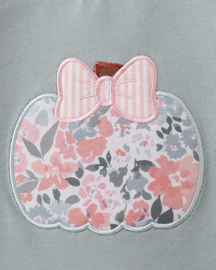Close-up of a light sage green garment featuring a scalloped applique patch shaped like a pumpkin, decorated with a pink and white striped bow and a pink, gray, and white floral pattern.