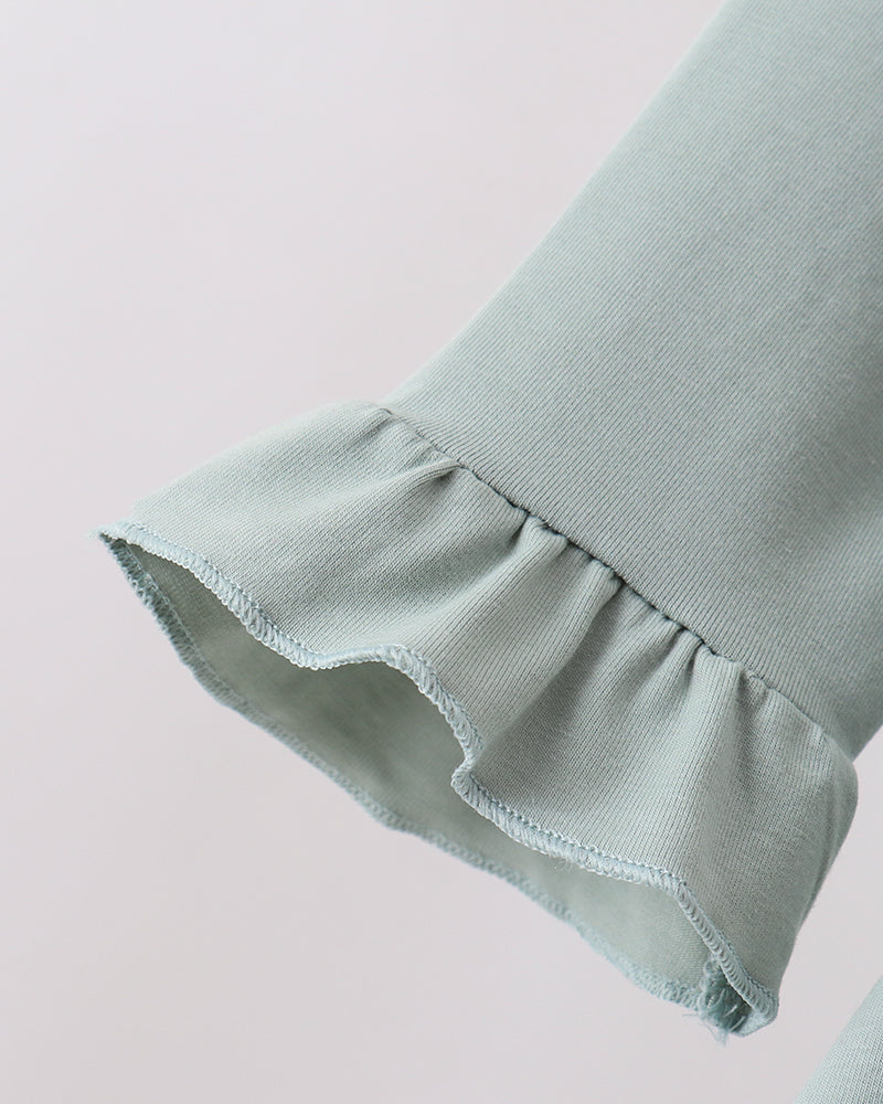 Close-up of a light sage green fabric garment with a ruffled cuff, showing the texture and hem detail against a plain light background.