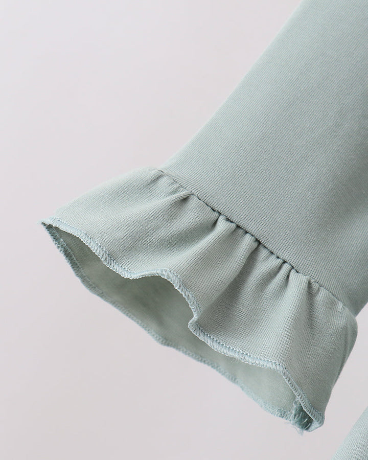 Close-up of a light sage green fabric garment with a ruffled cuff, showing the texture and hem detail against a plain light background.