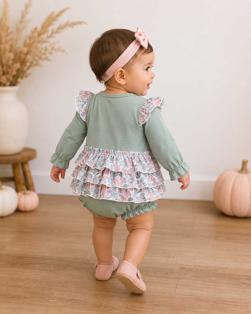 A toddler girl is shown from behind wearing a sage green long-sleeve romper with a floral tiered ruffle waist and matching green bloomers, along with a pink bow headband and pink shoes, in a decorated indoor room with pumpkins.