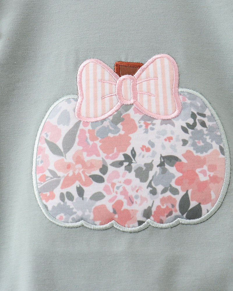 Close-up of a light sage green garment featuring a scalloped applique patch shaped like a pumpkin. The patch has a pink and white striped bow on top and a pink, gray, and white floral pattern below.
