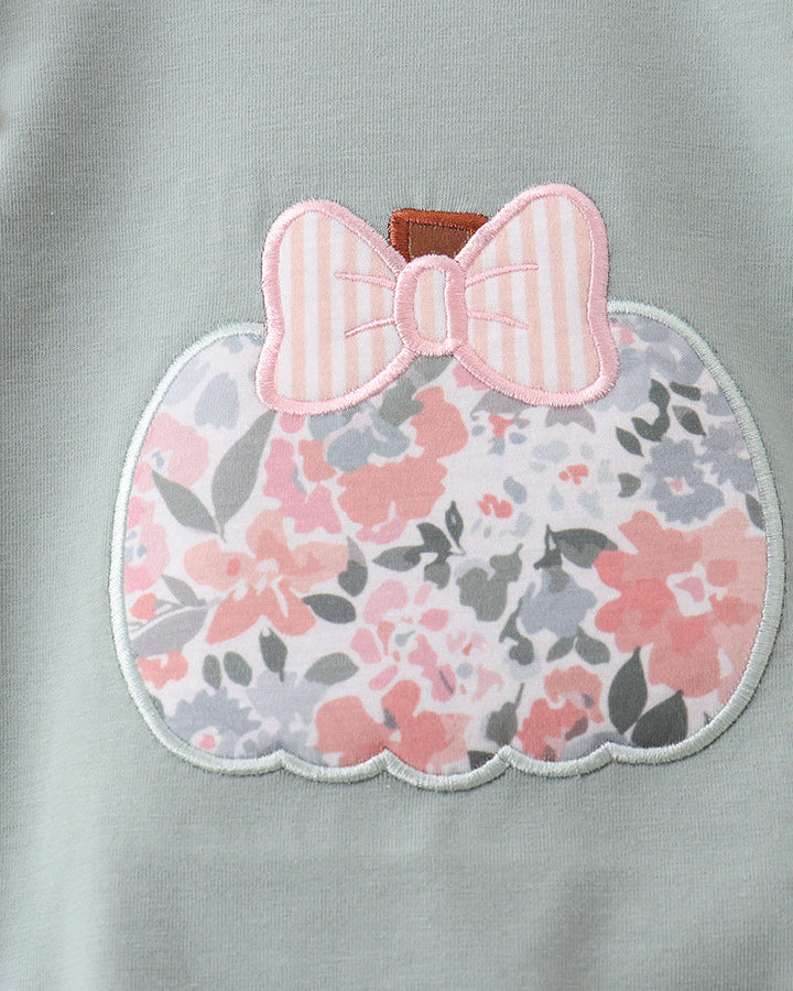 Close-up of a light sage green garment featuring a scalloped applique patch shaped like a pumpkin. The patch has a pink and white striped bow on top and a pink, gray, and white floral pattern below.