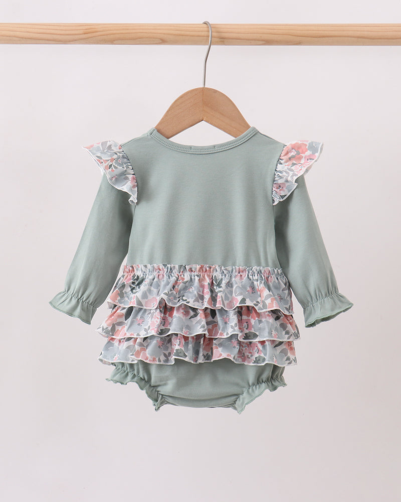 A baby long-sleeve romper in sage green, featuring ruffled floral-patterned shoulder straps and a tiered ruffle hem, hanging on a wooden hanger against a plain light background.