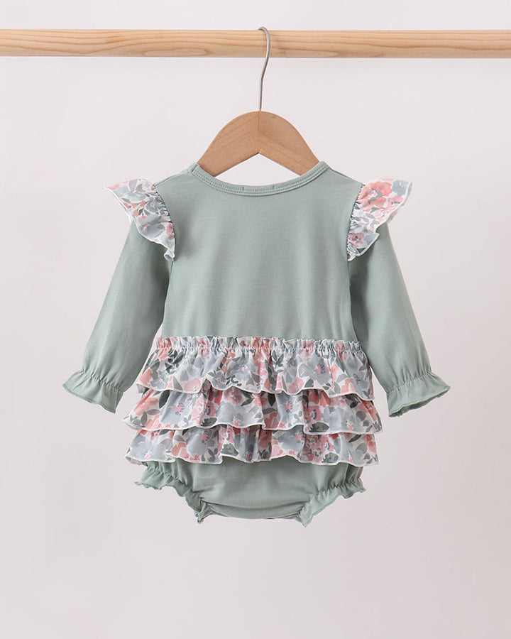 A baby long-sleeve romper in sage green, featuring ruffled floral-patterned shoulder straps and a tiered ruffle hem, hanging on a wooden hanger against a plain light background.