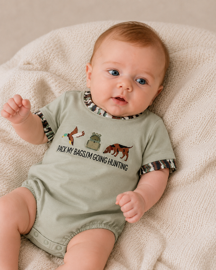 A baby lying on a textured cream blanket, wearing a sage green short-sleeve bodysuit with a camouflage-patterned collar and cuffs, featuring a hunting-themed graphic and the text &