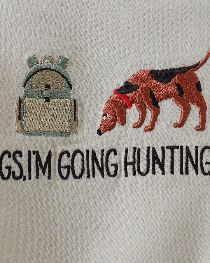 Close-up of a light beige embroidered graphic on a fabric surface, showing a backpack on the left, a brown dog with black patches and a red collar on the right, and partial black text reading &