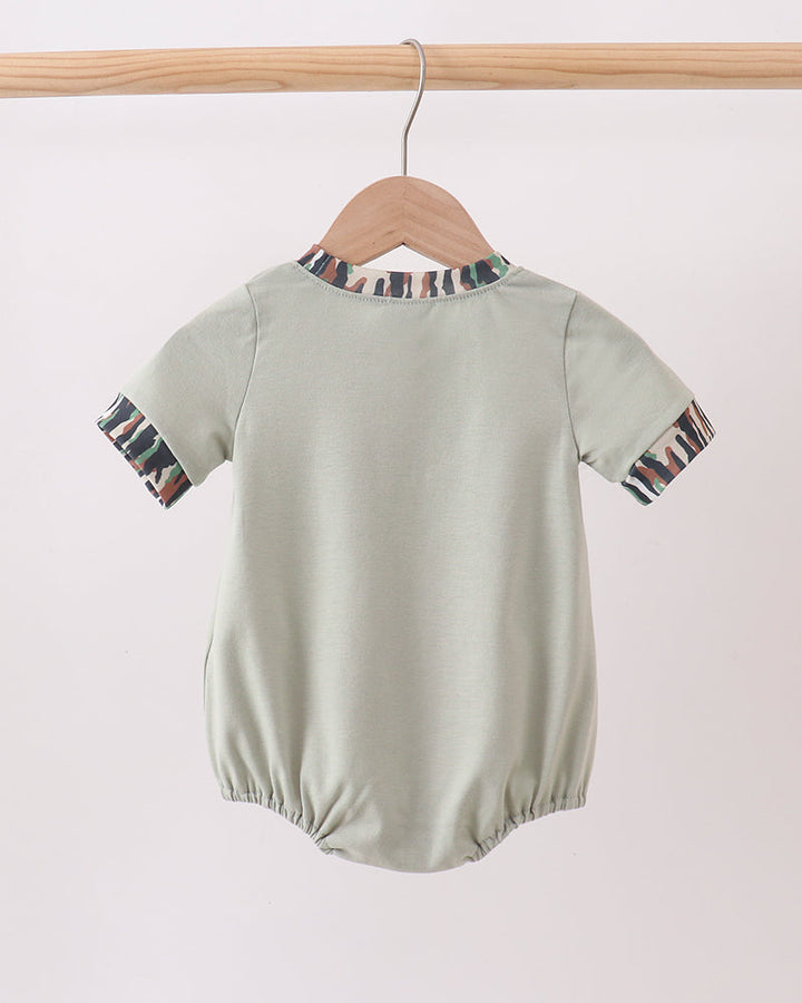 A sage green short-sleeve baby romper hanging on a wooden hanger from a wooden clothes rack, shown from the back. The romper features patterned trim along the neckline and sleeve cuffs.