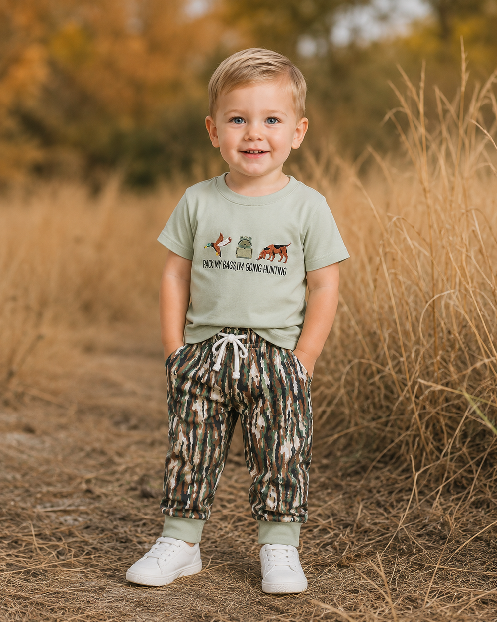 A young child standing outdoors in a field of dry grass, wearing a light sage green t-shirt with a hunting graphic and text, paired with camouflage patterned pants and white sneakers.