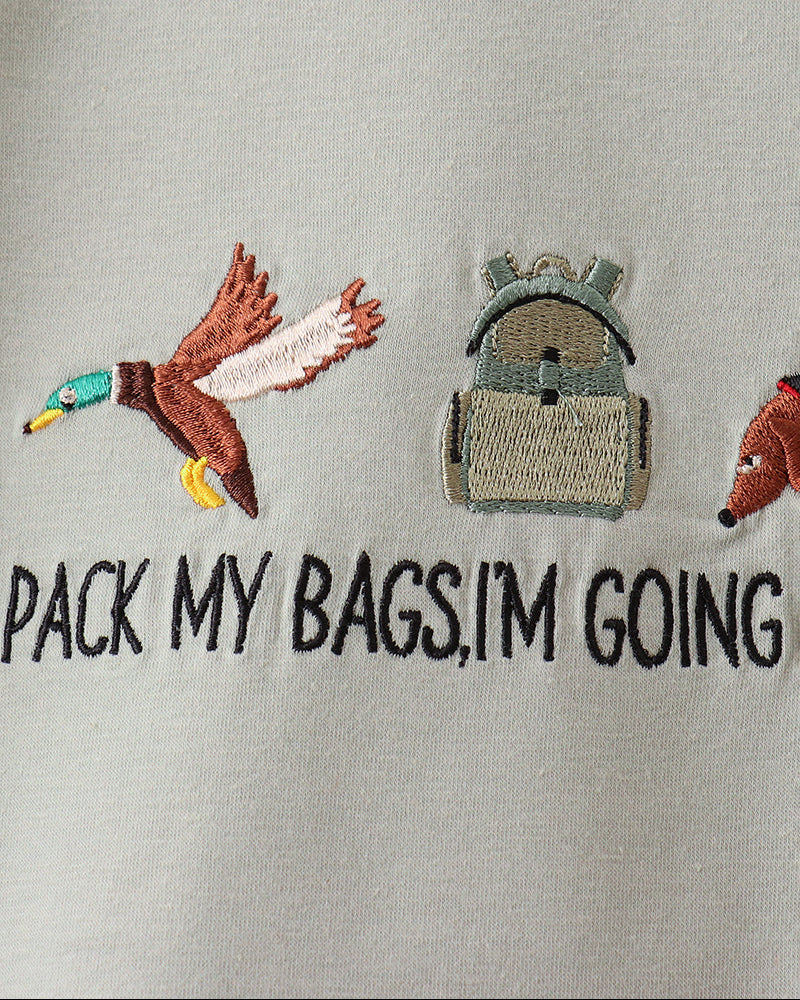 Close-up of a light gray or off-white fabric surface with embroidered graphics and text. Visible details include a brown-and-teal duck, a gray backpack, part of a fox, and the phrase &