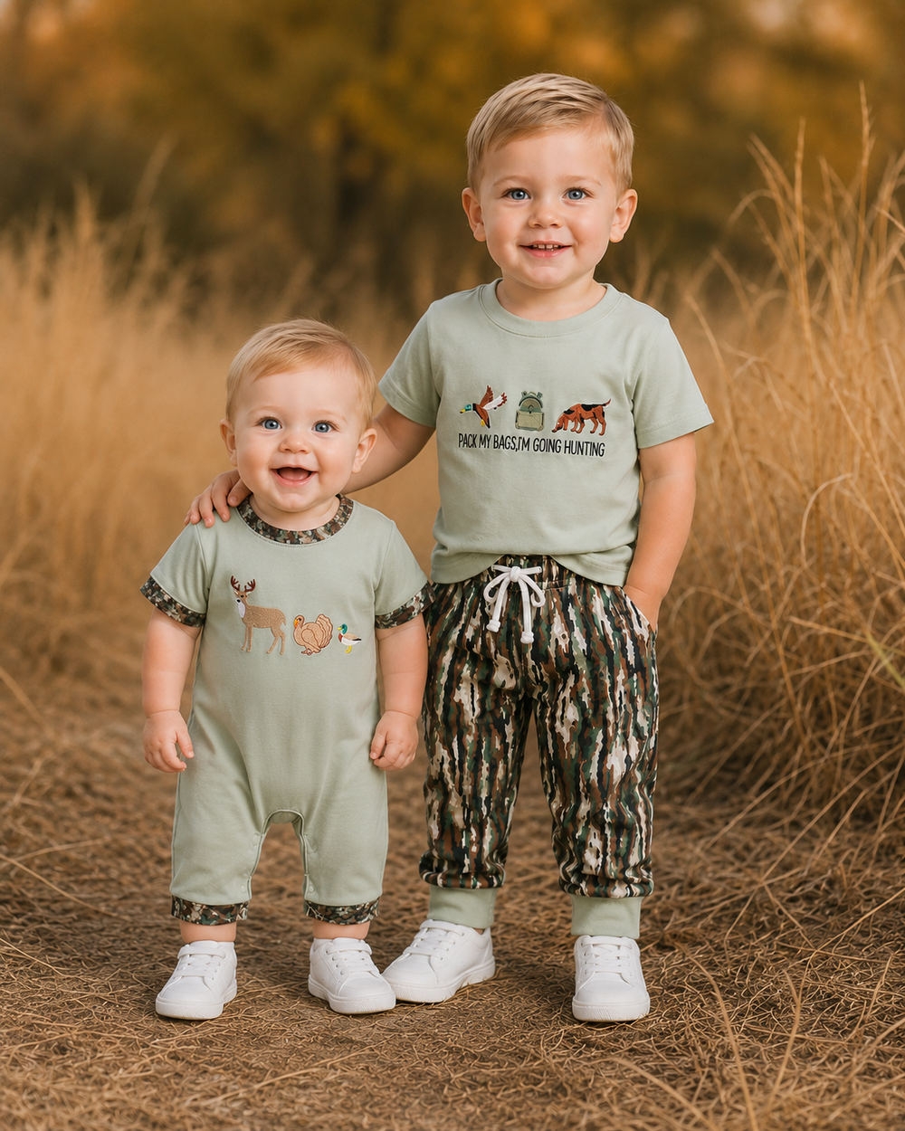 Two young children standing outdoors in a field of dry grass, wearing matching sage green t-shirts and camouflage-print pants. The older child has a graphic of a deer, fox, and the words 'THREE BOWS OF CROW HUNTING' on the front, and the younger child has a matching deer graphic on the front.