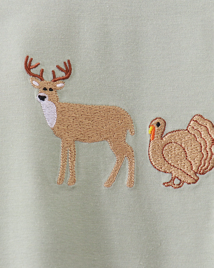 Close-up of a light sage green fabric featuring embroidered animals, including a tan deer with brown antlers and a matching tan turkey.