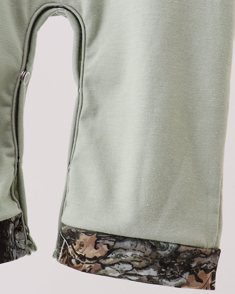 Close-up of a light sage green garment, likely a pair of pants or shorts, showing a curved leg opening and a contrasting camouflage-patterned hemband in brown, beige, and gray.