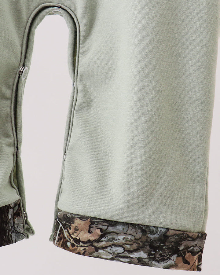Close-up of a light sage green garment, likely a pair of pants or shorts, showing a curved leg opening and a contrasting camouflage-patterned hemband in brown, beige, and gray.