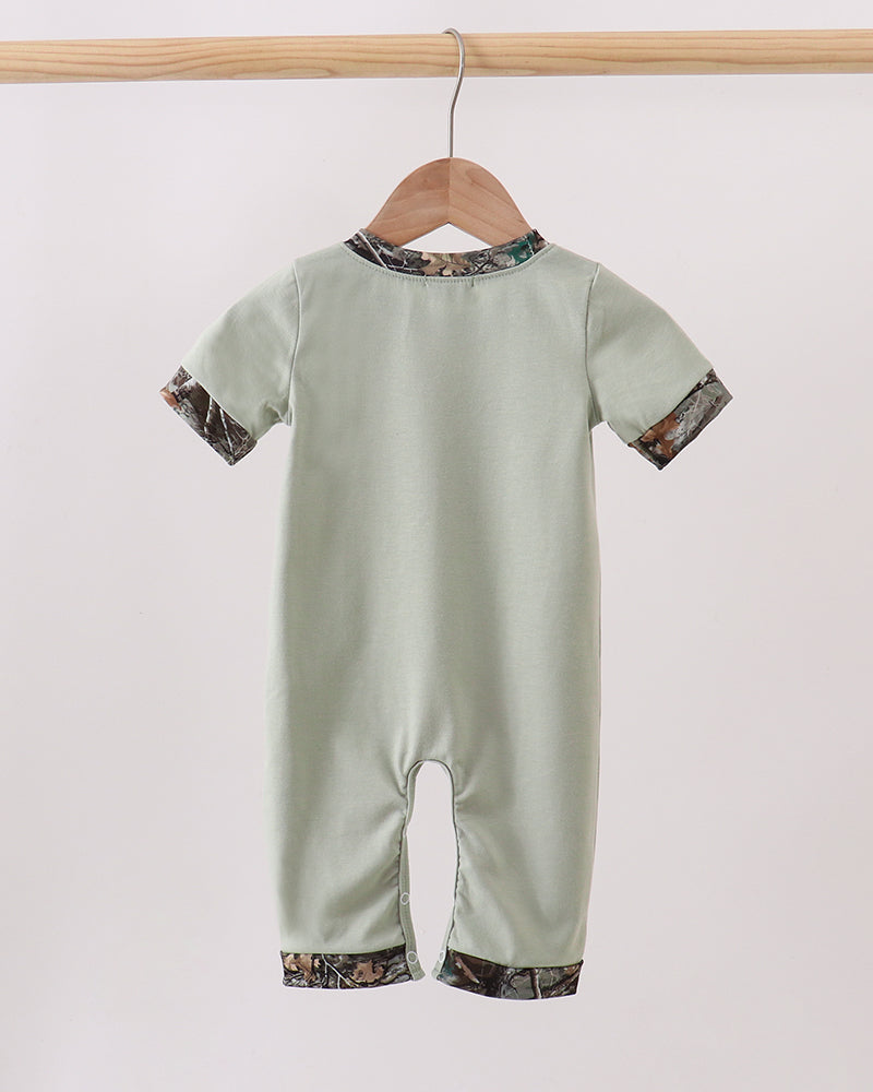 A baby short-sleeve romper in sage green, hanging on a wooden hanger from a light wood rod. The romper features a camouflage print trim along the collar, cuffs, and hems.