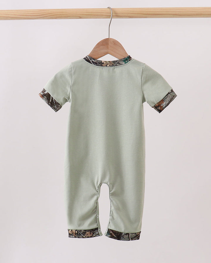 A baby short-sleeve romper in sage green, hanging on a wooden hanger from a light wood rod. The romper features a camouflage print trim along the collar, cuffs, and hems.
