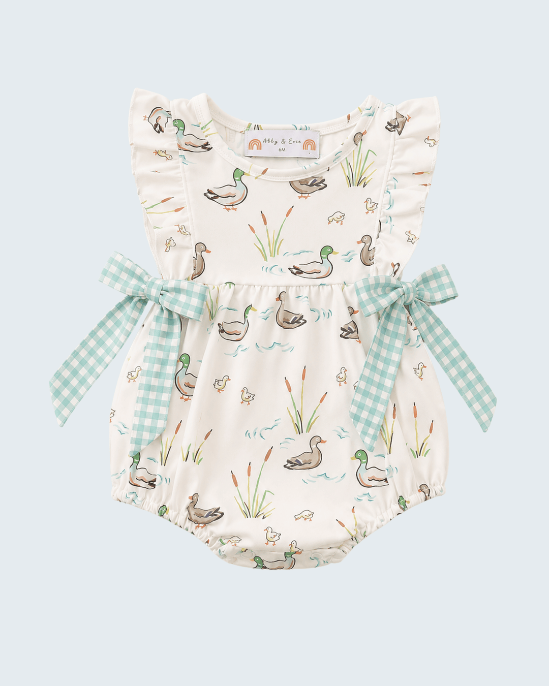 A baby romper in cream with a duck and reeds print, featuring ruffled shoulder straps, a matching gingham bow at the waist, and a white neck label reading &