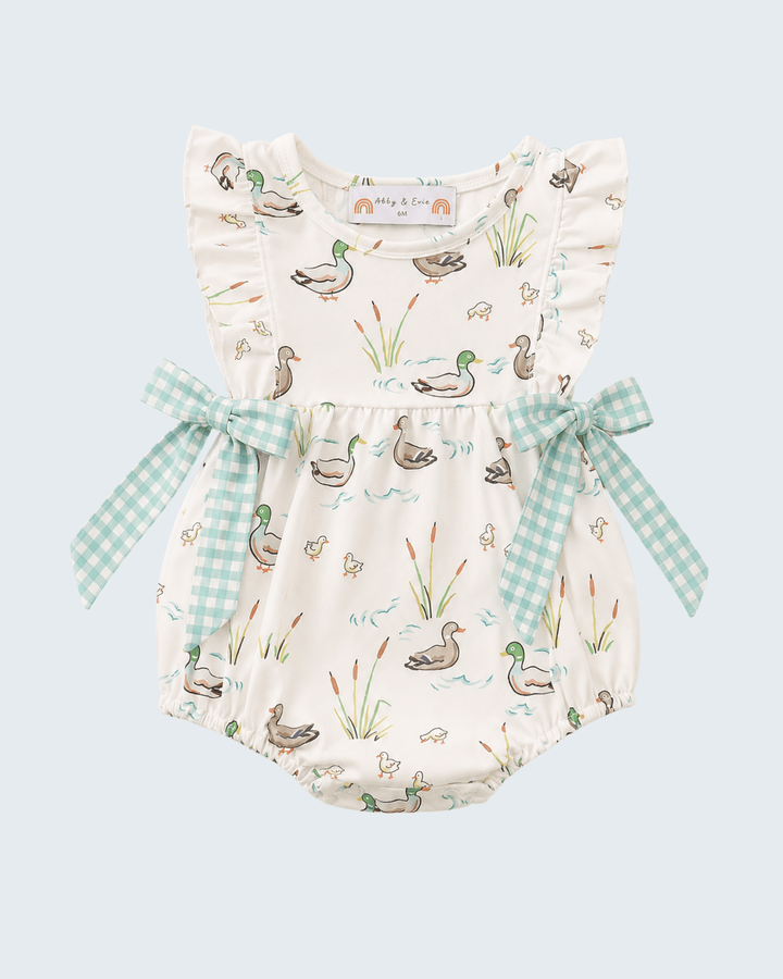 A baby romper in cream with a duck and reeds print, featuring ruffled shoulder straps, a matching gingham bow at the waist, and a white neck label reading &