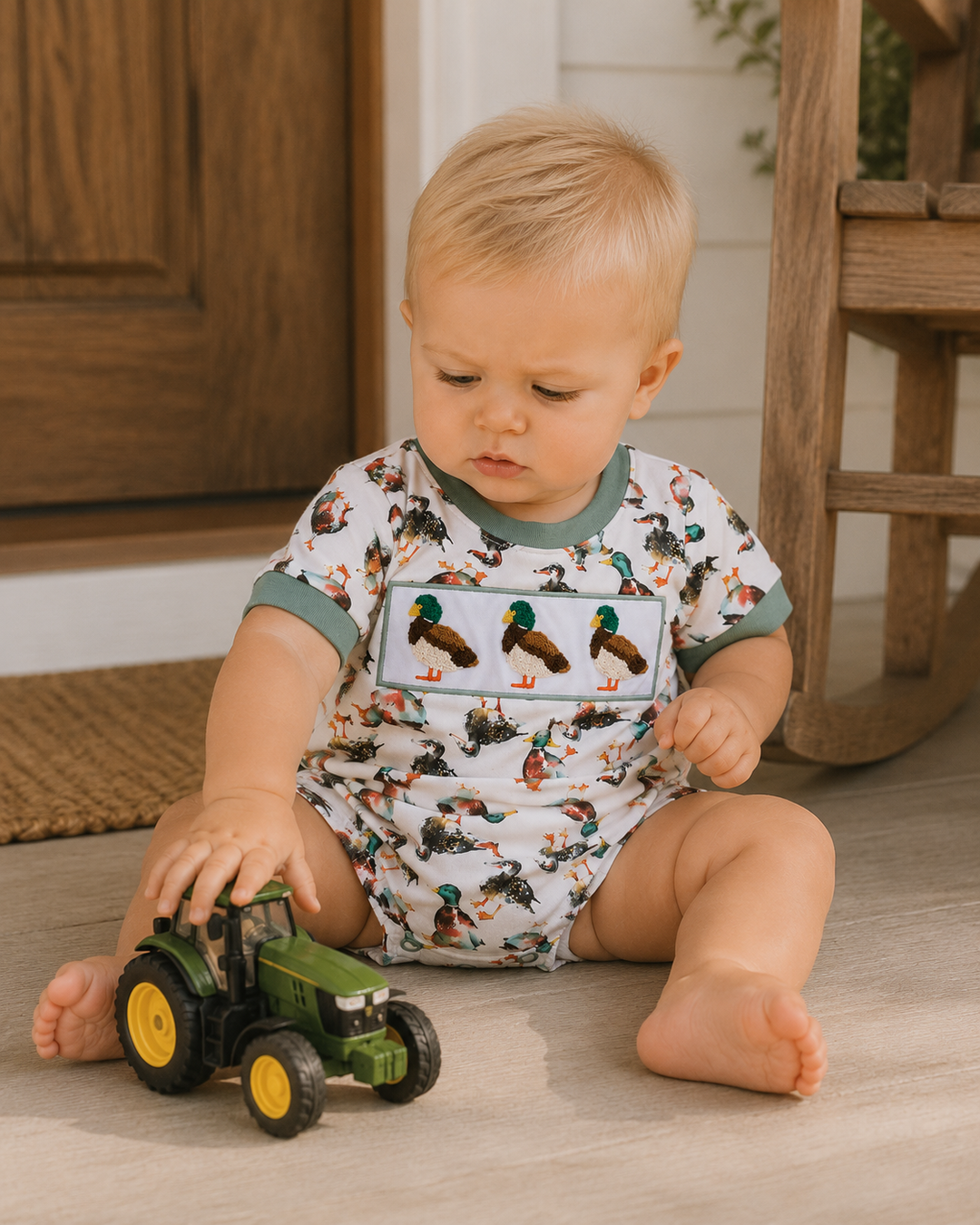 A baby sitting on a light wood floor playing with a green toy tractor, wearing a white short-sleeve romper printed with ducks and a large duck graphic on the chest, with green trim.