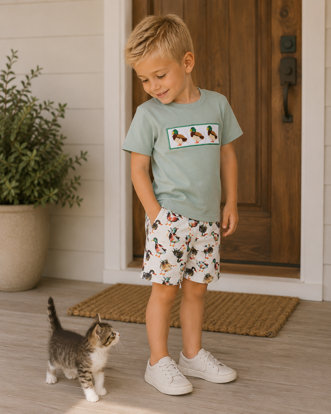 A young boy wearing a sage green t-shirt with a chicken graphic and white shorts with a black and red chicken print, standing on a wooden porch next to a small tabby cat.
