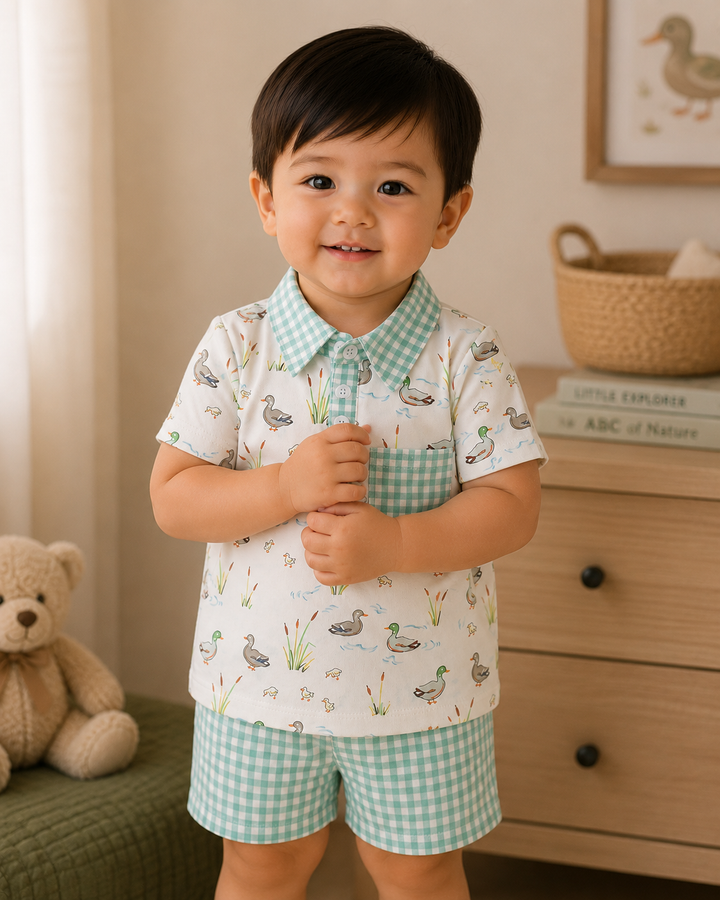 A smiling toddler boy wearing a two-piece matching set with a short-sleeve collared shirt and shorts. The set features a white base with a light green gingham pattern on the collar, pocket, and shorts, and a print of ducks, grass, and water lilies.