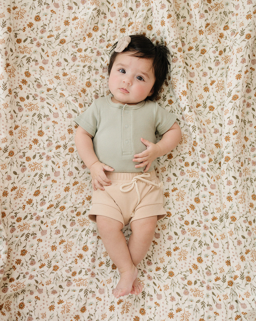 Baby in green top and beige shorts lying on a floral-patterned blanket