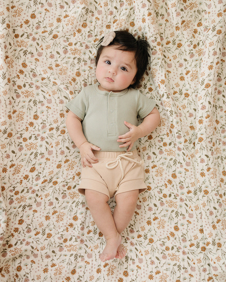 Baby in green top and beige shorts lying on a floral-patterned blanket