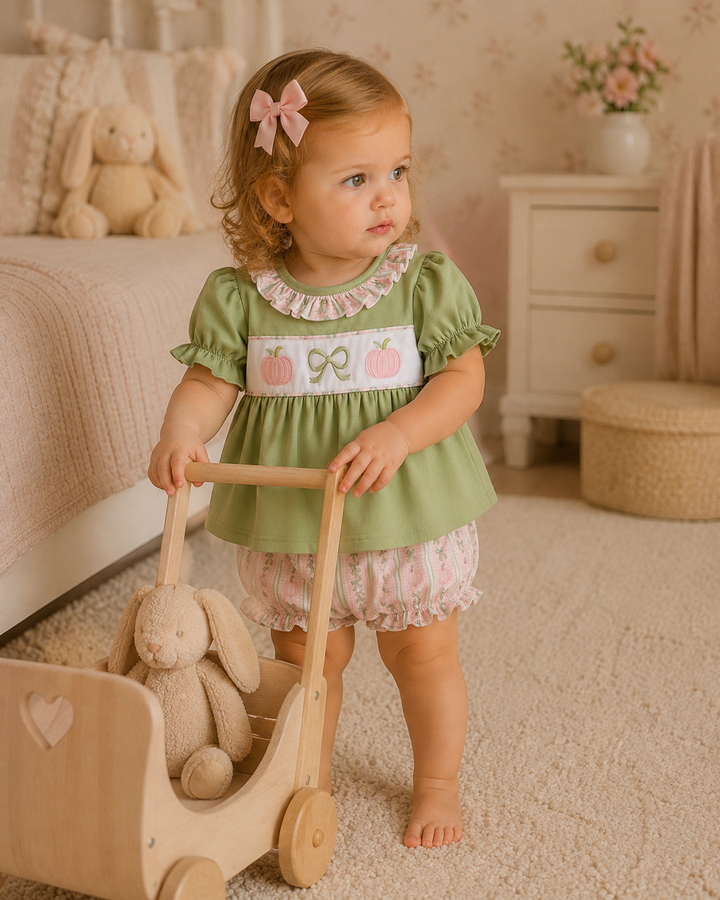 A toddler girl in a sage green dress with a white embroidered chest panel stands in a nursery setting, pushing a wooden toy cart with a stuffed bunny inside. The room has a beige carpet, a white nightstand, and a teddy rabbit on the bed.