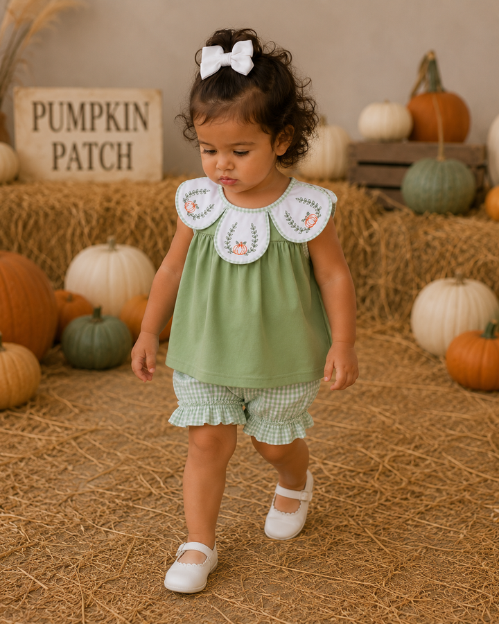 A toddler girl walking in a fall-themed studio setting with hay, pumpkins, and a sign that reads &