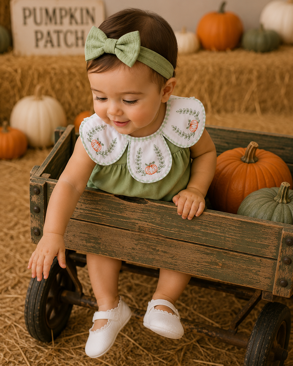 A baby girl wearing a sage green dress with a white embroidered Peter Pan collar and a matching green bow headband, sitting in a rustic wooden wagon decorated with pumpkins on a hay-covered floor.