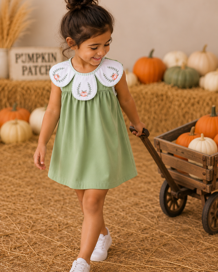 A young girl wearing a sage green dress with a white Peter Pan collar featuring embroidered floral details, walking in a fall-themed pumpkin patch setting with straw and pumpkins.