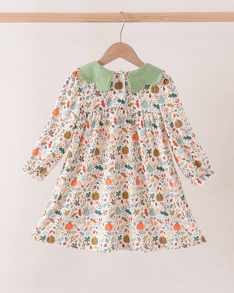 A long-sleeve baby dress with a green collar and an all-over autumn pumpkin and leaf print, hanging on a wooden hanger against a plain light beige wall.