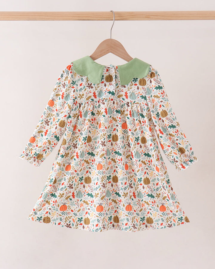 A long-sleeve baby dress with a green collar and an all-over autumn pumpkin and leaf print, hanging on a wooden hanger against a plain light beige wall.