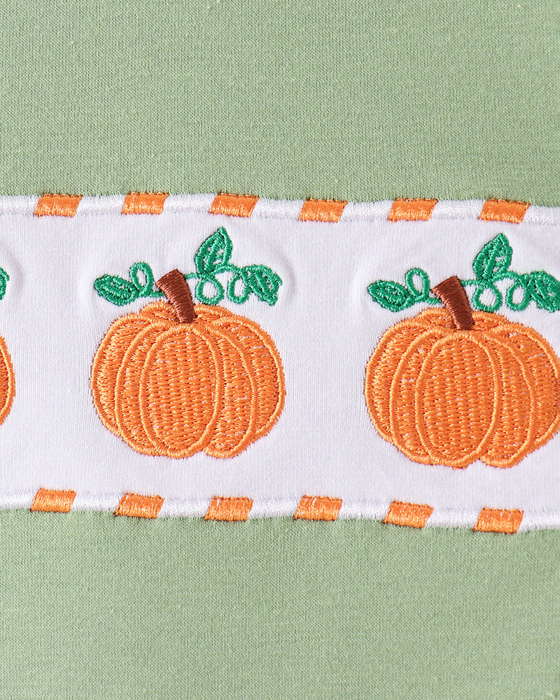 A close-up of a white embroidered trim featuring orange pumpkins with green leaves and brown stems, bordered by orange and white stripes, set against a light green fabric background.