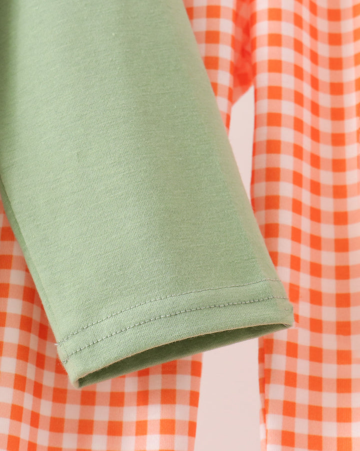 Close-up of a sage green sleeve with a neat hem, layered over an orange and white gingham patterned fabric.