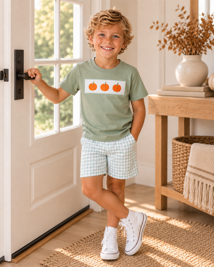 Young boy standing by a outside door. He is wearing a pumpkin shorts outfit.