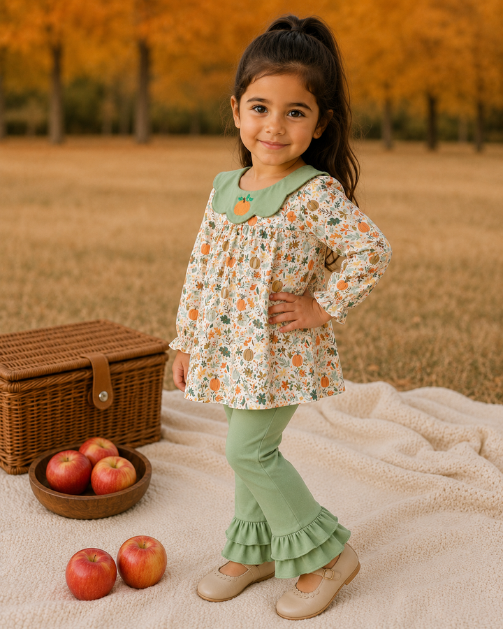 A young girl stands outdoors on a cream blanket with a wicker picnic basket and apples nearby. She is wearing a long-sleeve cream blouse with a green floral print and a green Peter Pan collar decorated with a small orange pumpkin motif, paired with sage green ruffled pants and beige flats.