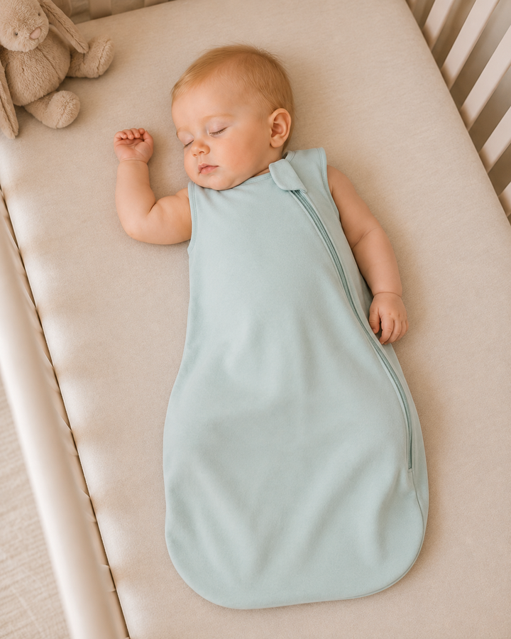 A sleeping baby lying on their side on a cream-colored crib mattress, wearing a sleeveless, light blue sleep sack with a zipper closure.