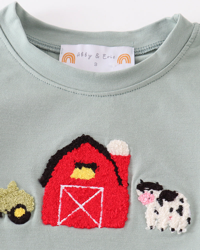 A close-up of a light sage green baby t-shirt featuring embroidered farm animals. The design includes a red barn with white trim, a black and white cow with a pink nose, and a small yellow tractor on the left.