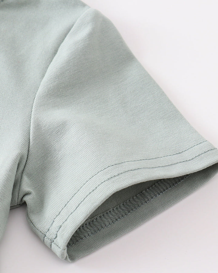 Close-up of a light sage green fabric garment, showing the texture of the woven material and a rolled cuff with neat stitching, on a plain white background.