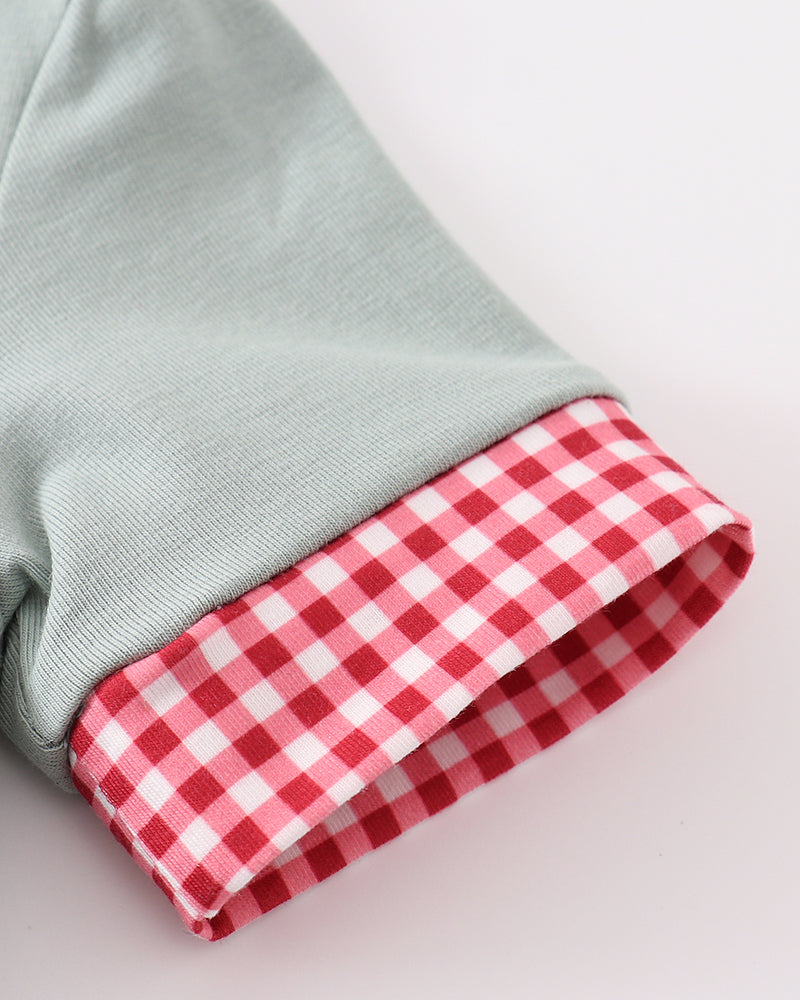 Close-up of a light sage green garment cuff turned up to show a red and white gingham checkered lining, on a plain light background.